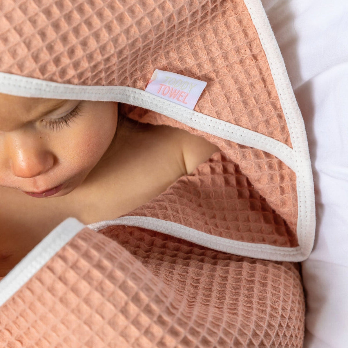 Child wrapped in a pink towel with a visible label, lying on a white surface.