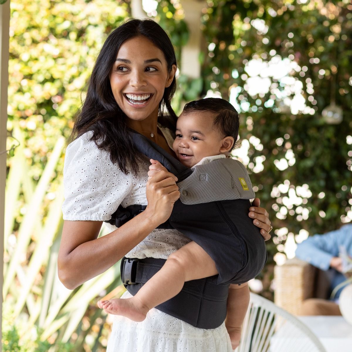 Woman holding a baby in a black carrier outdoors with greenery in the background