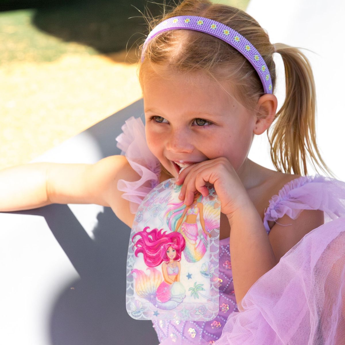 Young girl in a purple dress with a fairy-themed headband, sitting outdoors drinking from sinchie.