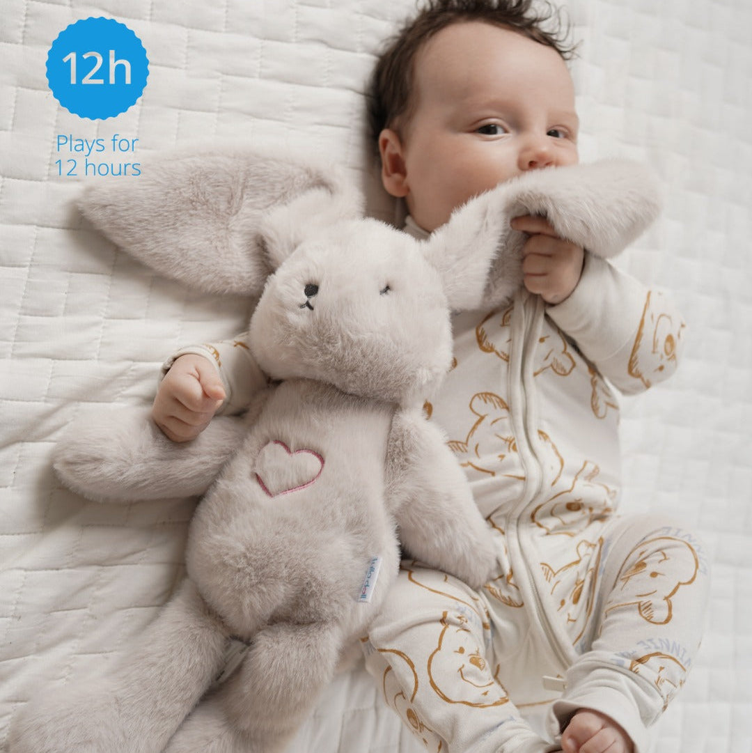 Baby lying on a bed with a plush bunny toy, wearing a patterned onesie.