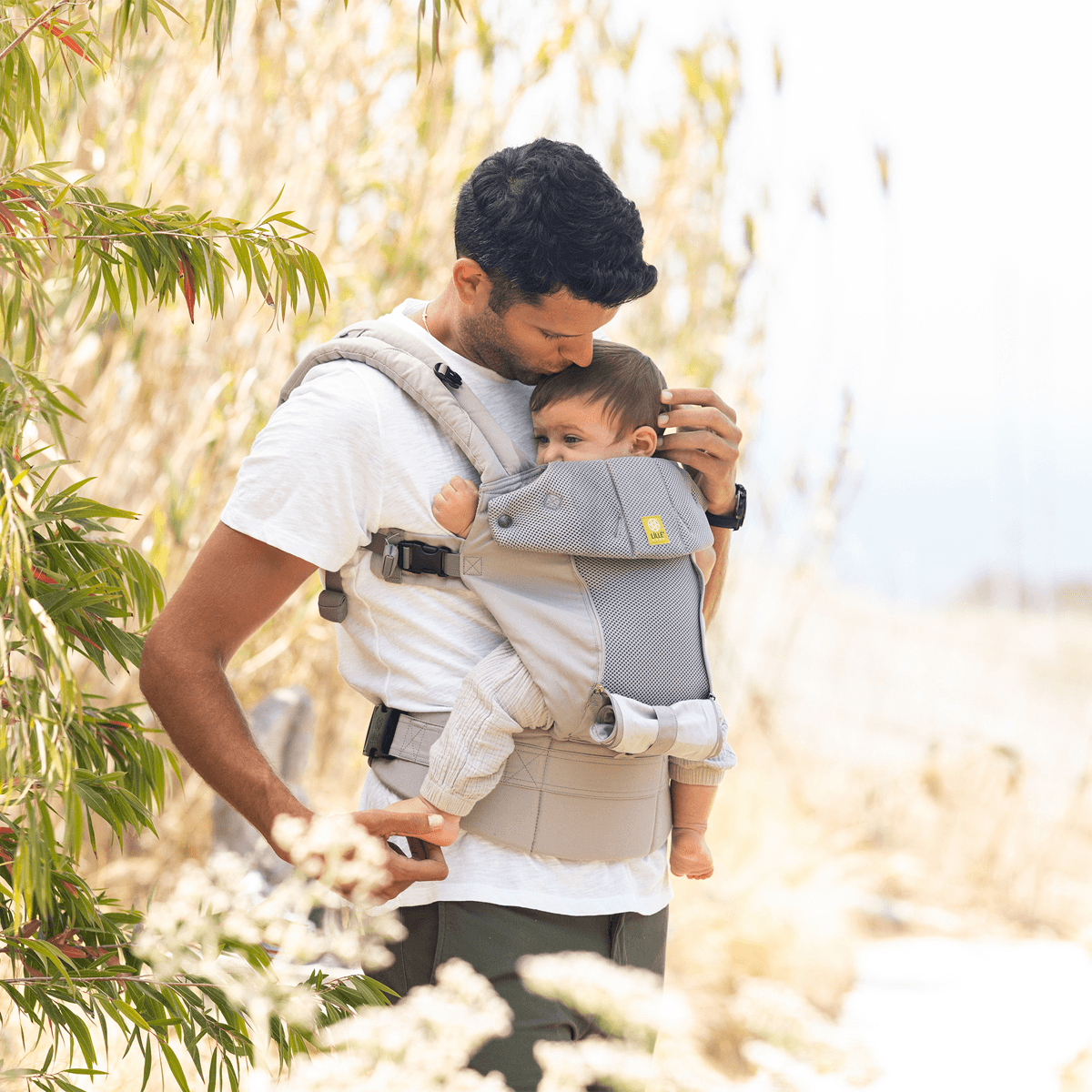 Man holding a child in a baby carrier outdoors with greenery in the background