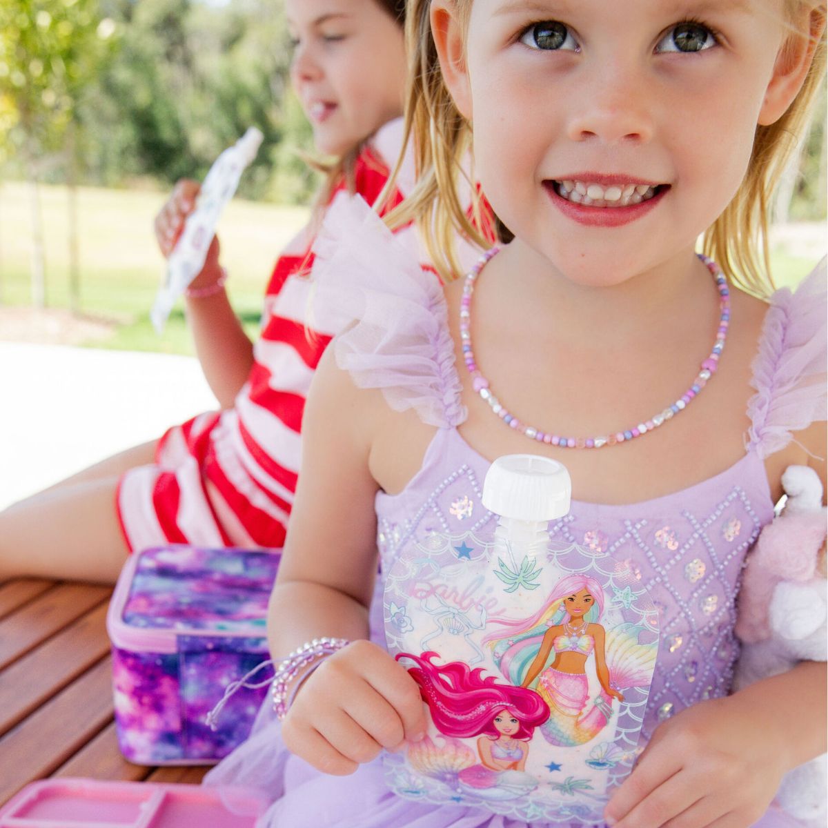 Child in a purple dress with a princess design, sitting outdoors with another child. Drinking a barbie themed sinchie.