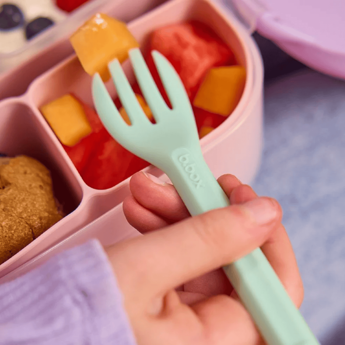 Hand holding a green fork over a pink bento box with compartments of food