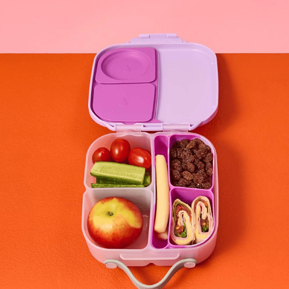 Pink bento box with compartments filled with fruits, vegetables, and snacks on a pink and orange background
