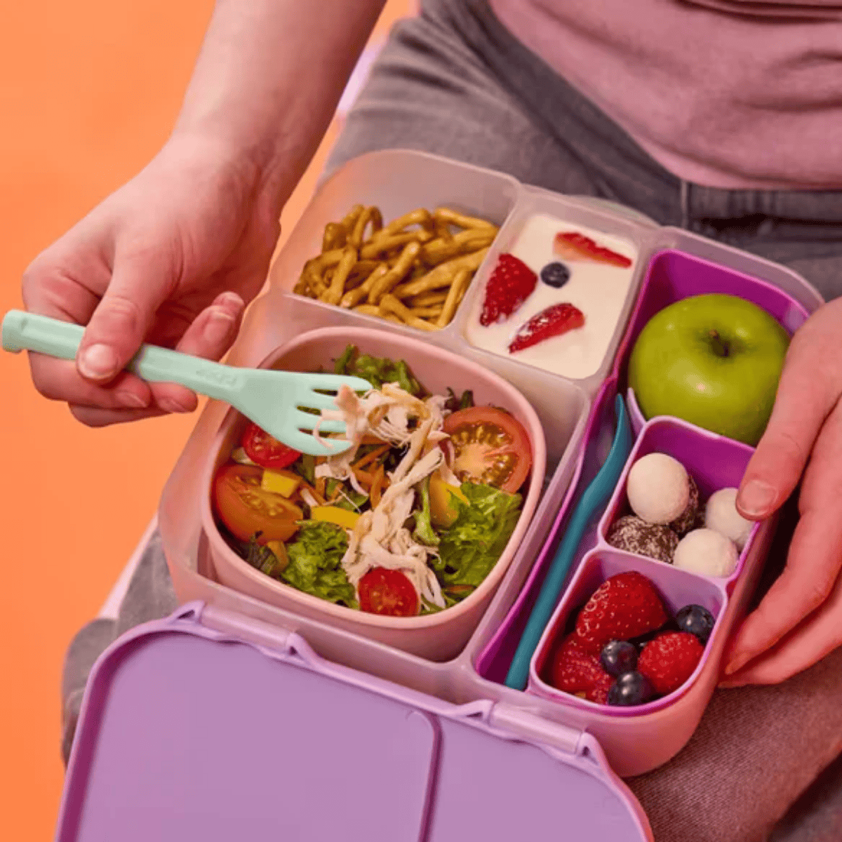 Person using a green spoon to eat from a colorful bento box filled with food on an orange background