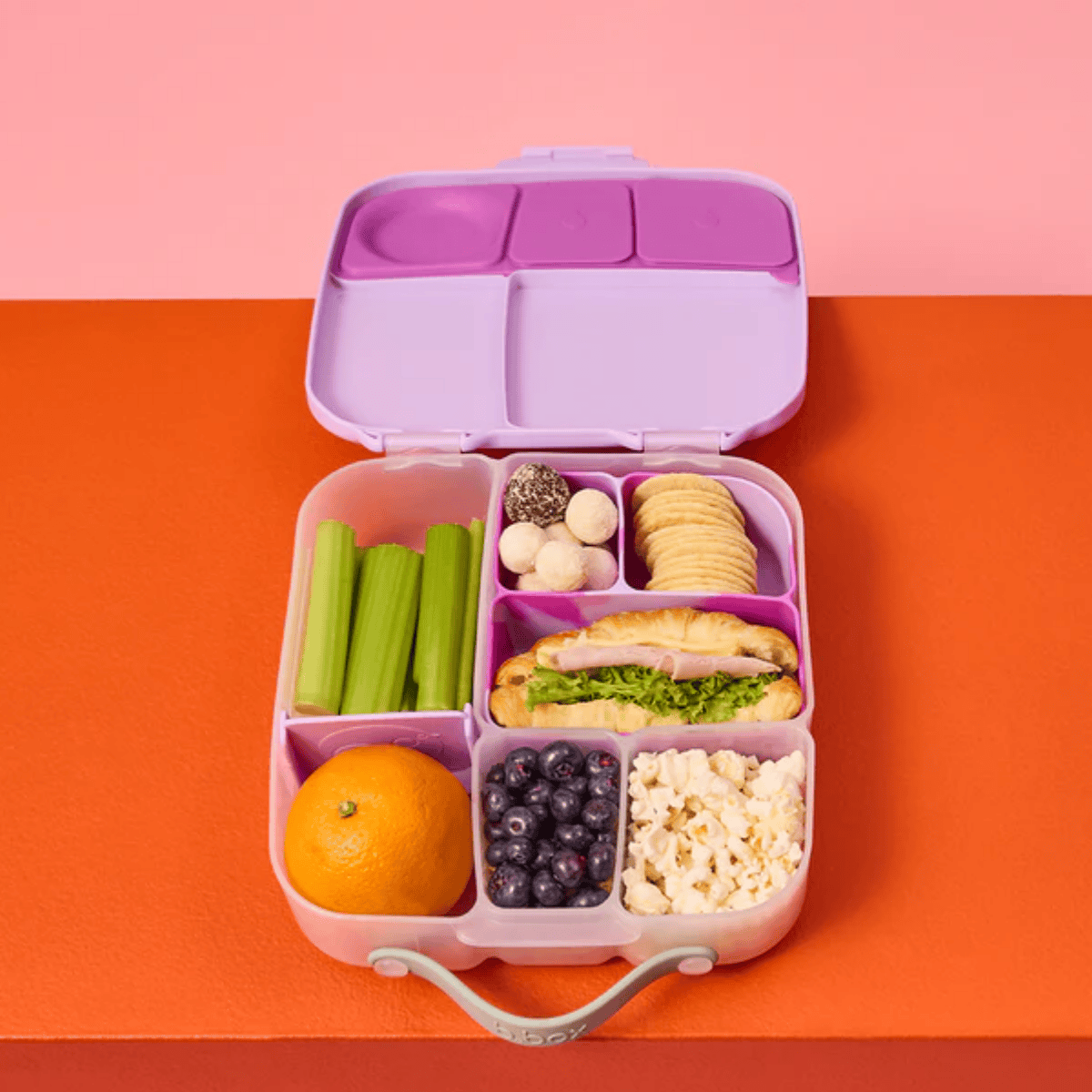 Purple bento box with compartments filled with various food items on a two-tone pink and orange background