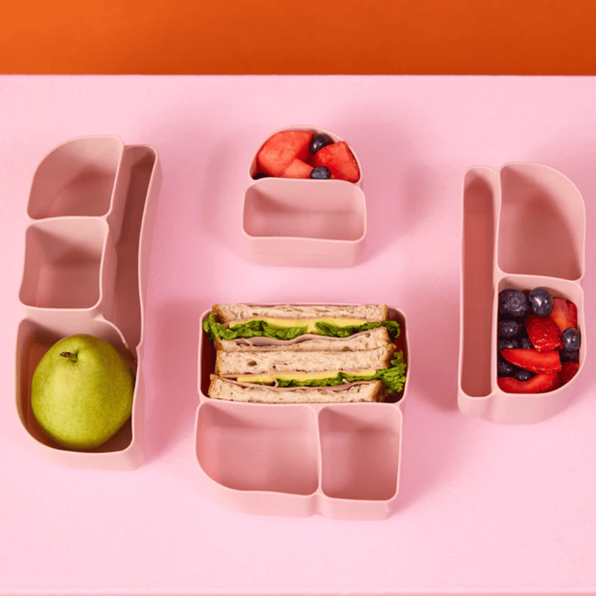 Pink bento box with compartments containing sandwiches, fruits, and berries on a pink background