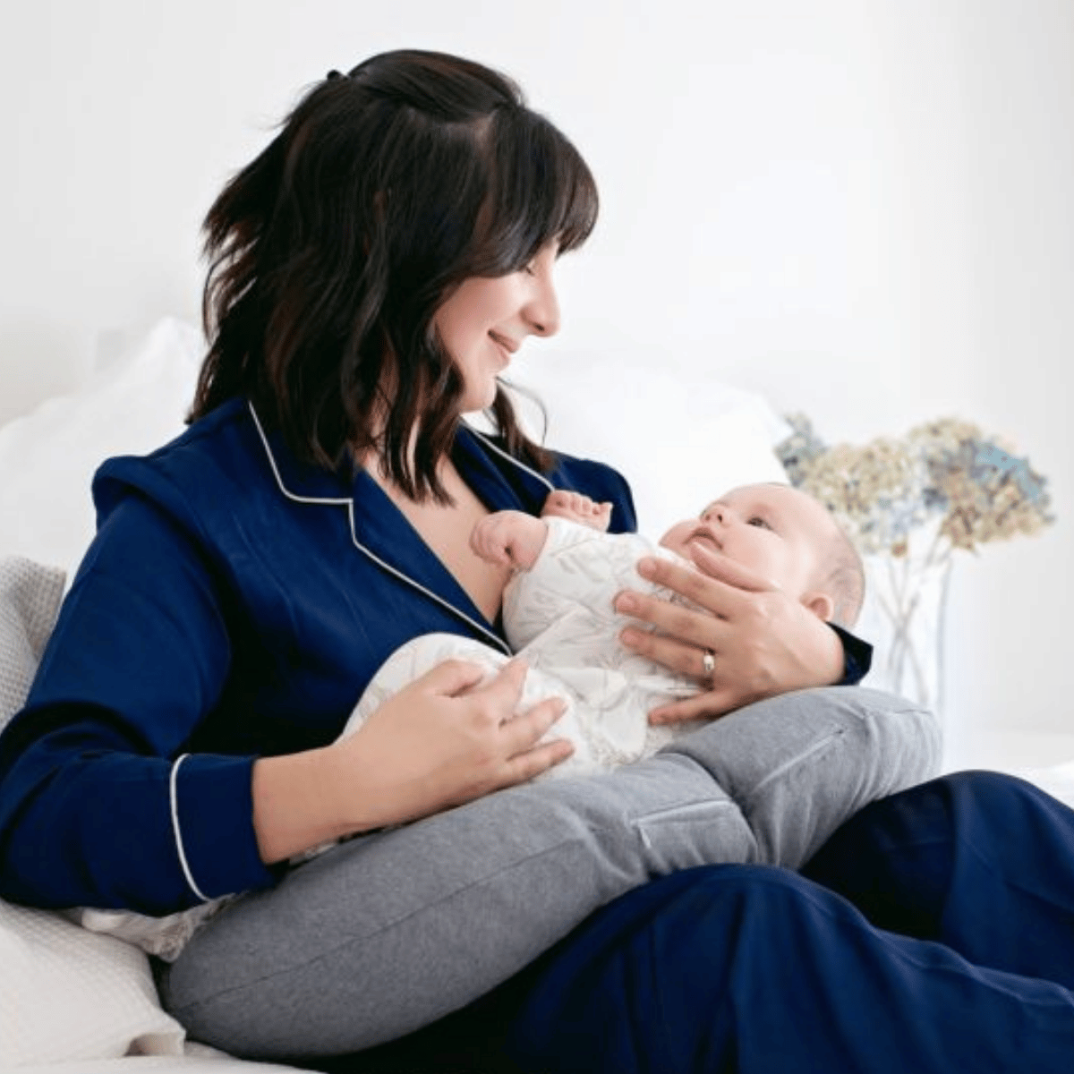 Woman in blue pajamas holding a baby on a bed surrounded by a breastfeeding cushion