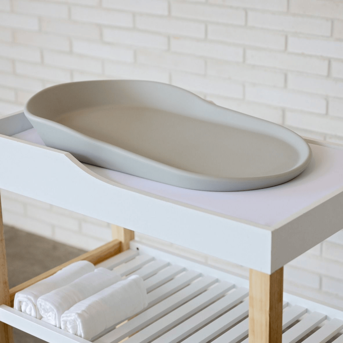 Gray oval change mat on a white countertop with a wooden shelf below against a white brick wall.
