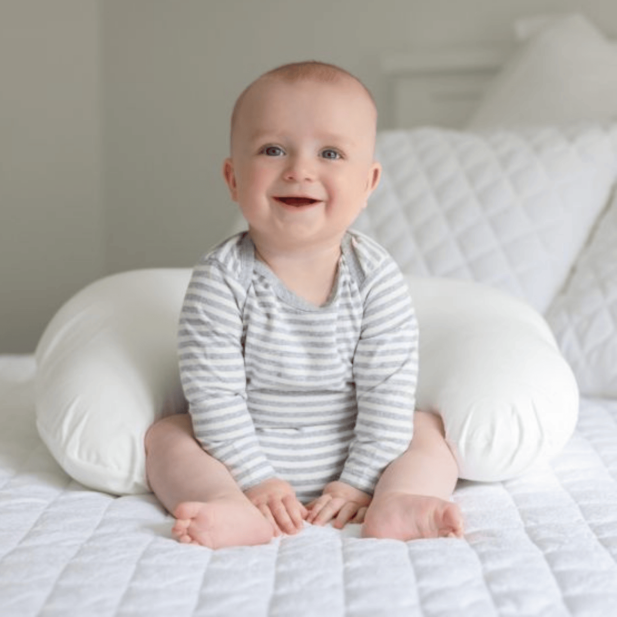 Baby sitting on a white pillow with a gray and white striped shirt, smiling.