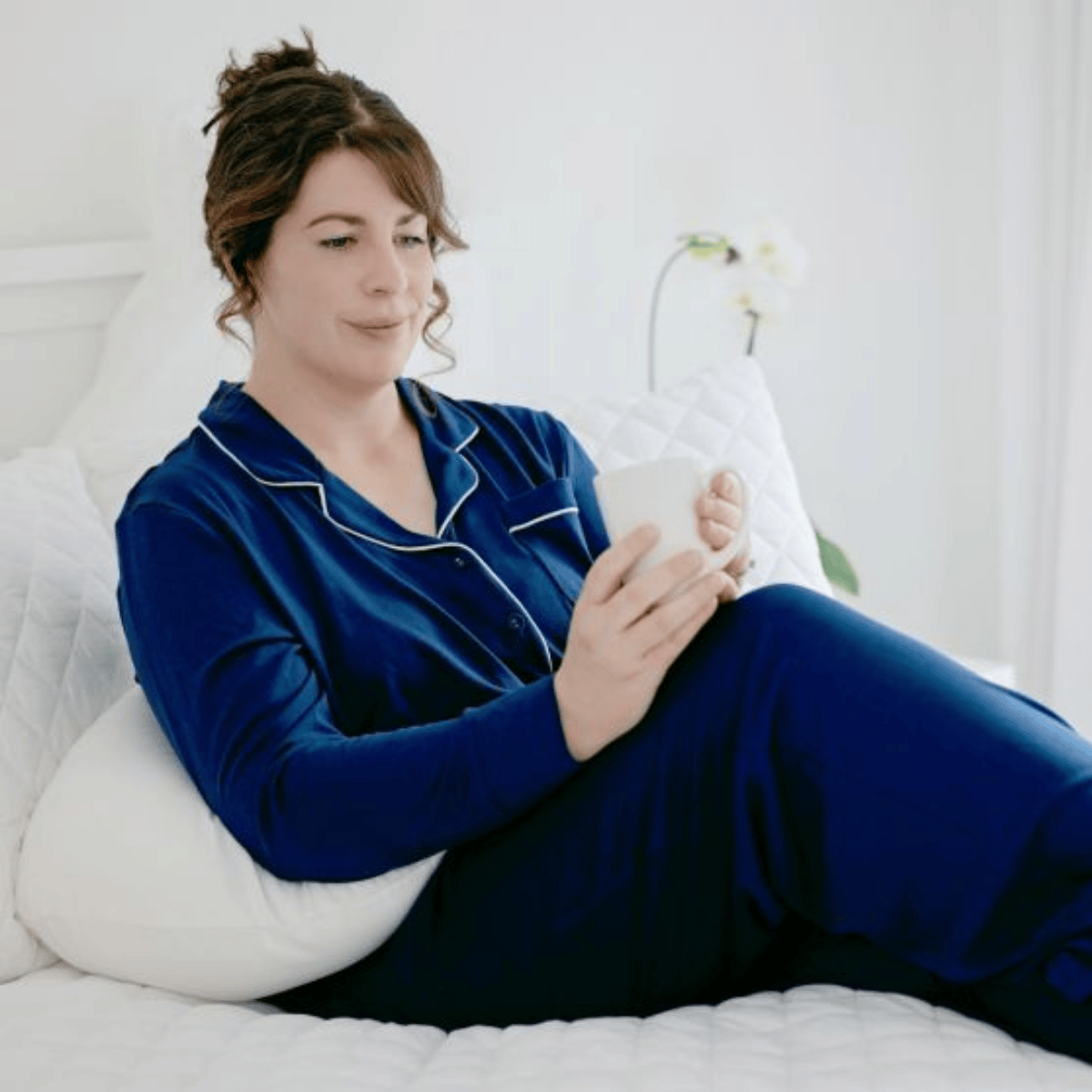 Woman in blue pajamas holding a white mug on a bed.