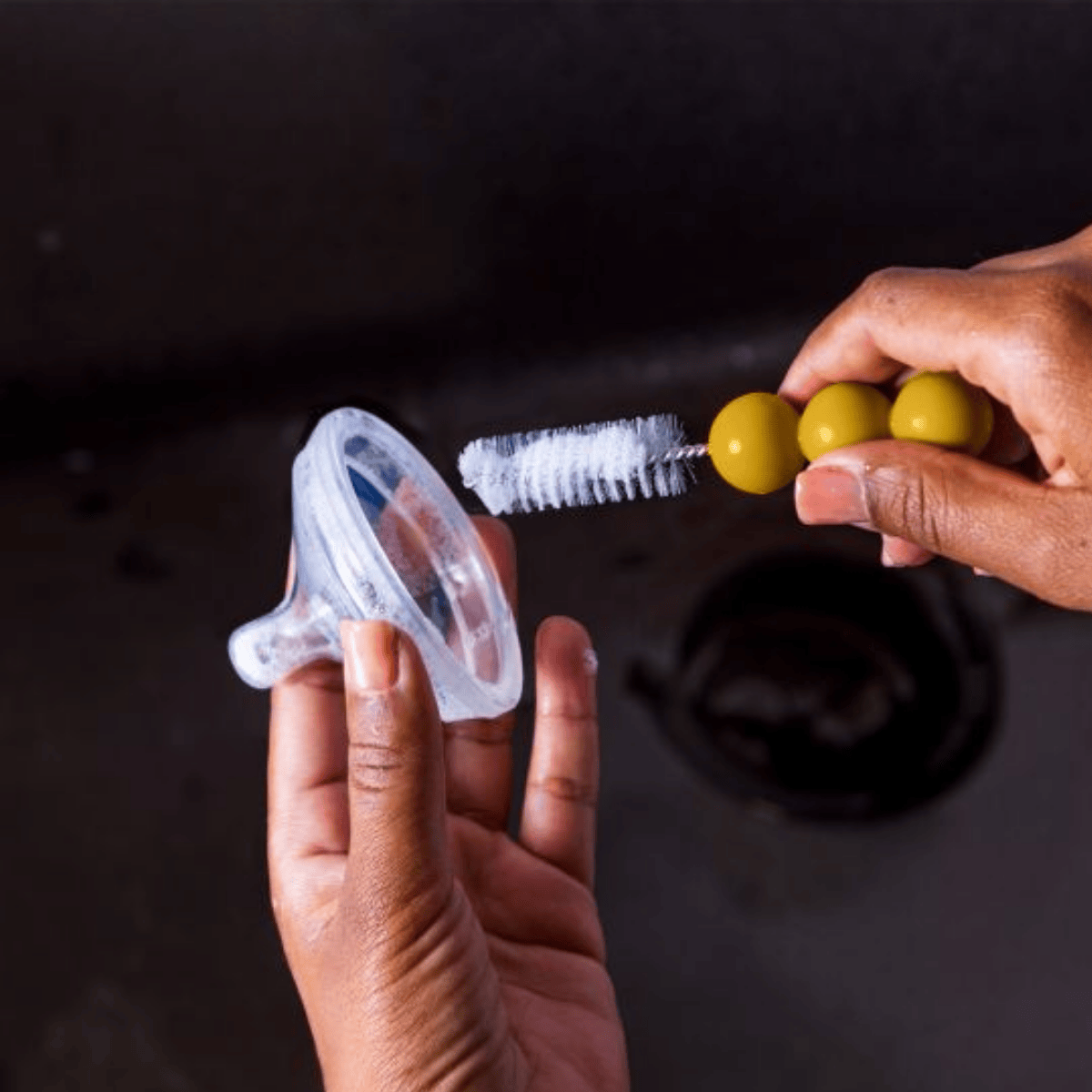 Hands holding a baby bottle nipple, brush, and green olives against a dark background