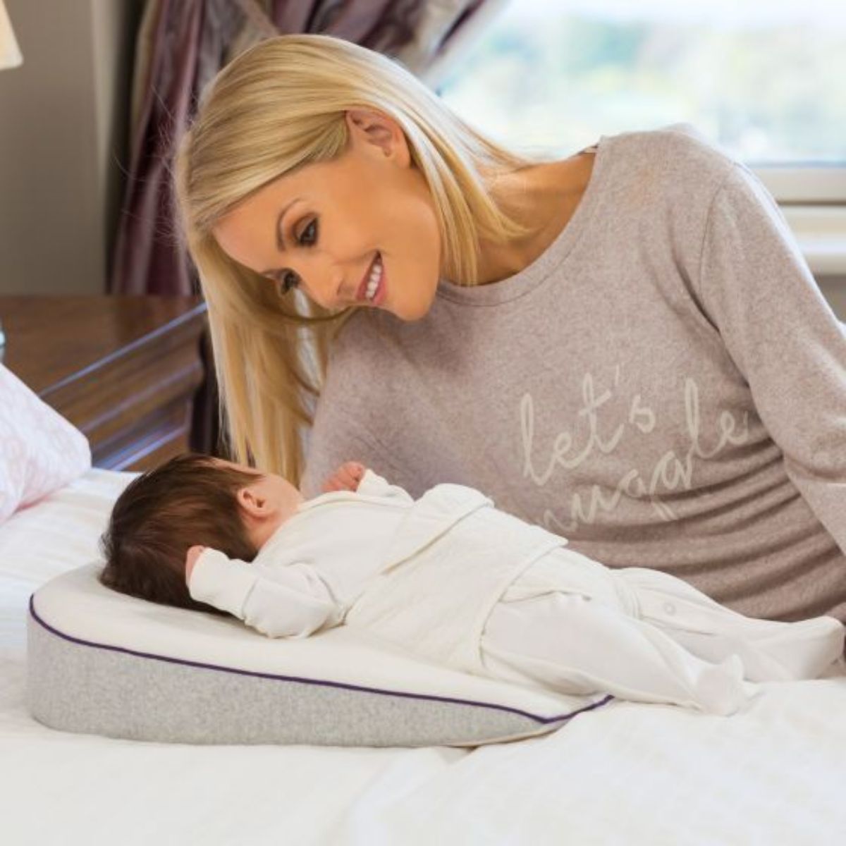 Woman in a gray sweatshirt with 'let's snuggle' text, leaning over a baby lying on a wedge-shaped pillow.