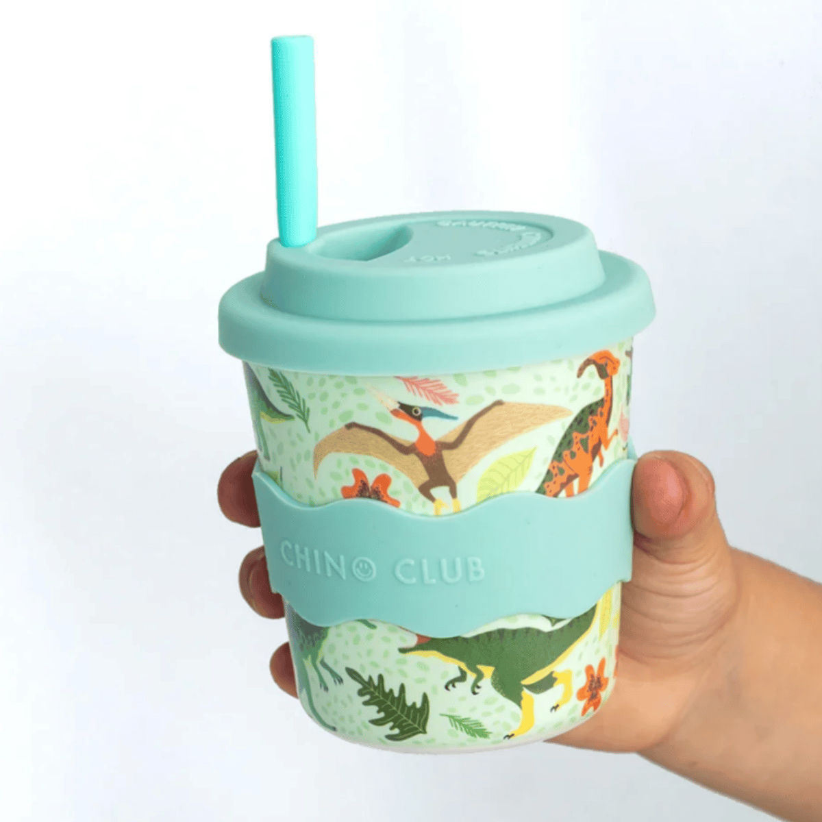 A hand holding a reusable tumbler cup with a light blue lid, straw, and silicone sleeve. The cup body has a dinosaur and plant pattern on a pale green background, with the text 'CHINO CLUB' embossed on the sleeve.
