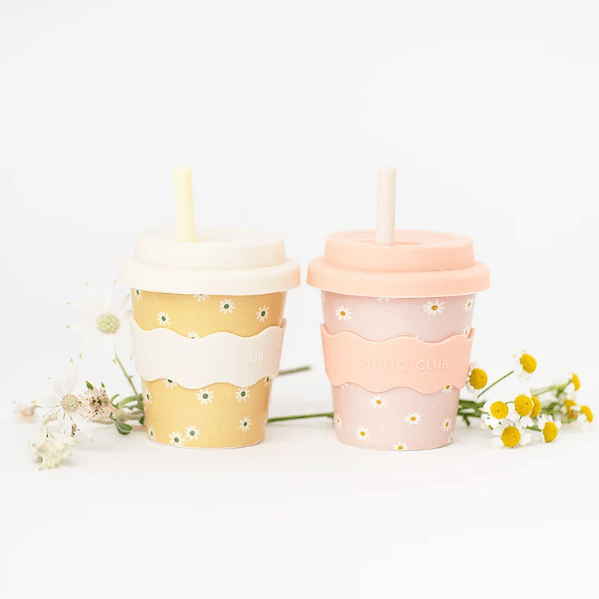 Two reusable paperless cups with lids and straws, decorated with small daisy flowers. One cup is yellow with a white band and yellow flower print, and the other is pink with a peach band and pink flower print.