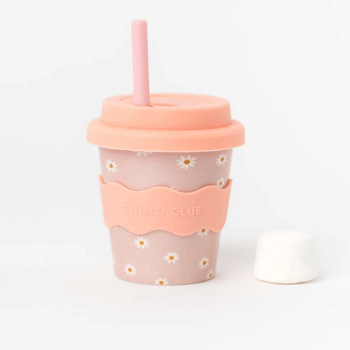 A pink reusable fluffy cup with a peach-colored silicone sleeve, lid, and straw, decorated with a white daisy pattern. The sleeve features the text 'CHINO CLUB'.