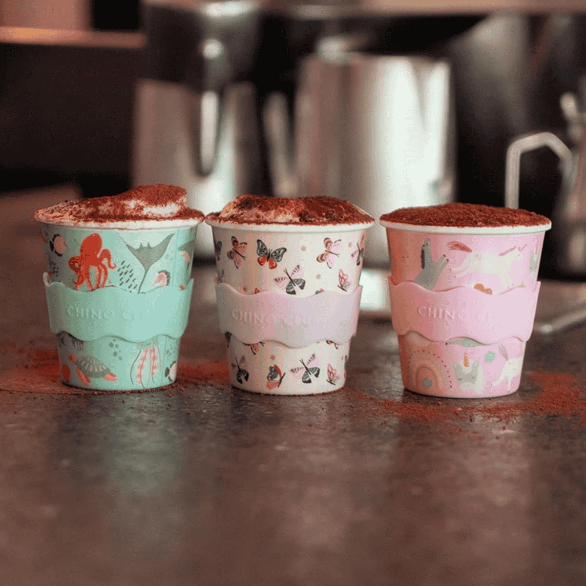 Three patterned paper fluffy cups with pastel sleeves sit on a countertop, each filled with a cocoa or coffee drink topped with cocoa powder. The cups have whimsical illustrations including sea life, butterflies, and unicorns, and the sleeves are branded CHINO CLIC.