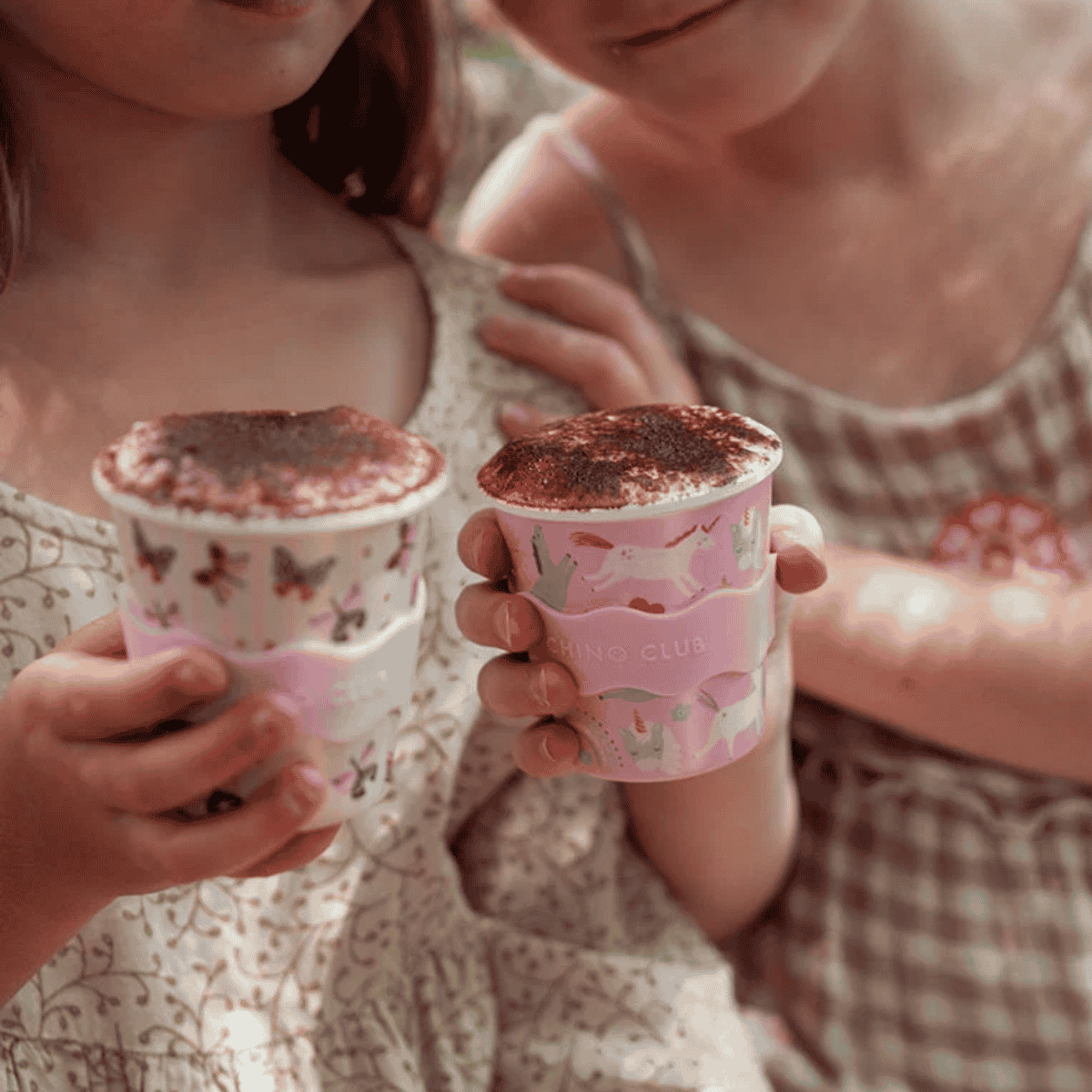 Two children in patterned dresses holding pink ceramic mugs filled with cocoa, with the brand name 'SHOOGH CLUB' visible on the mugs.