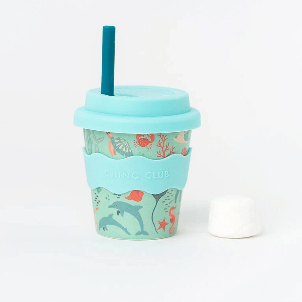 A reusable bamboo travel tumbler with a light blue lid and straw, featuring a teal silicone sleeve with an undersea-themed print of dolphins, starfish, and coral. A small white marshmallow is placed to the right on a clean white background.