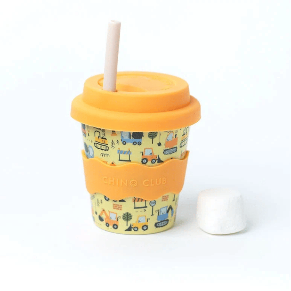A reusable paper fluffy cup with a yellow construction-themed print, an orange silicone sleeve and lid, a beige straw, and a white marshmallow resting beside it on a white background.