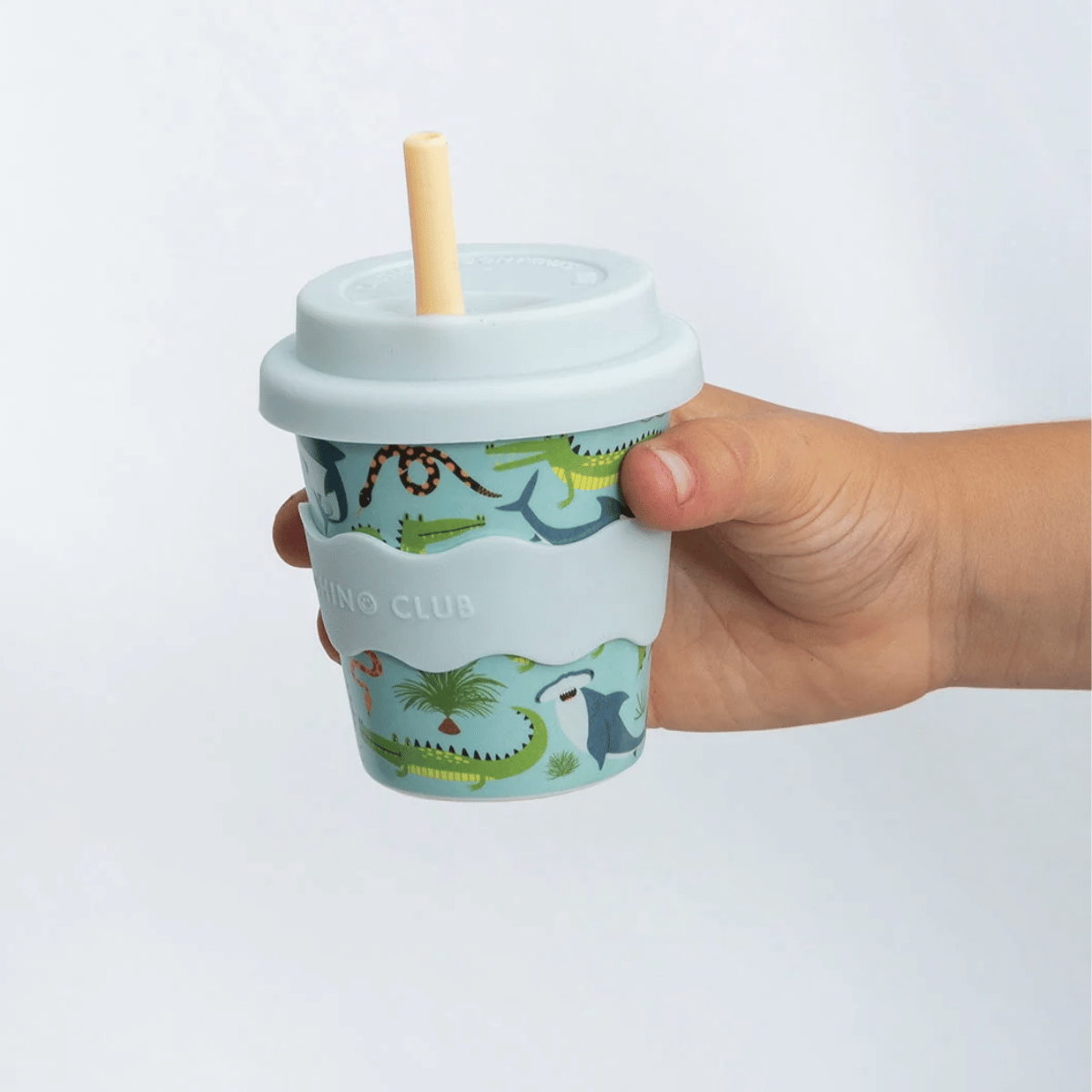 A hand holding a light blue reusable cup with a straw and lid. The cup features a colorful dinosaur-themed wrap design with green, blue, and yellow illustrations, and the words 'DINO CLUB' are printed on the sleeve.