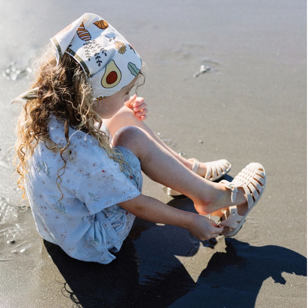 Child sitting on the beach wearing a patterned headband and sandals.