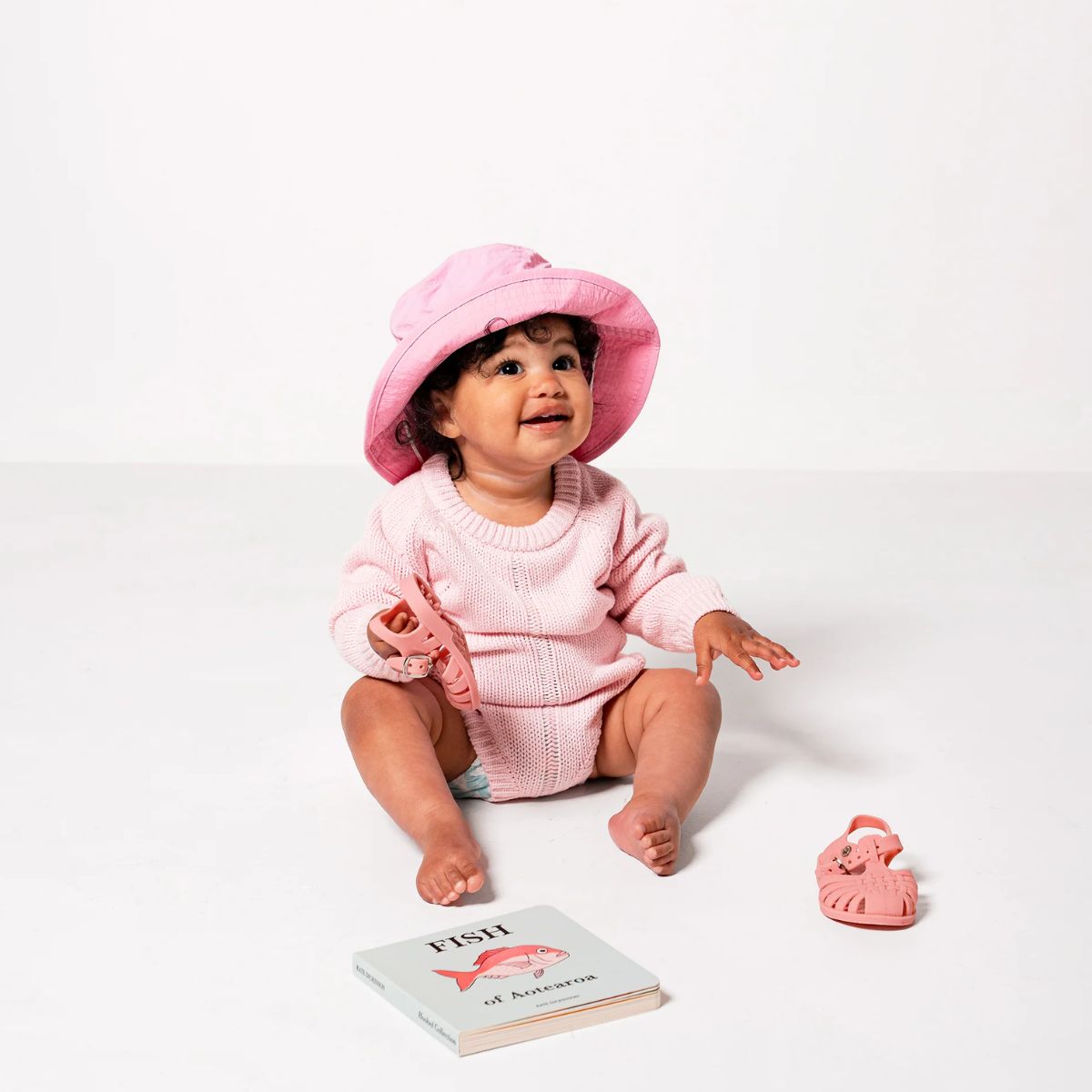 Baby in pink outfit with matching hat, sitting on a white surface with a book and toy.