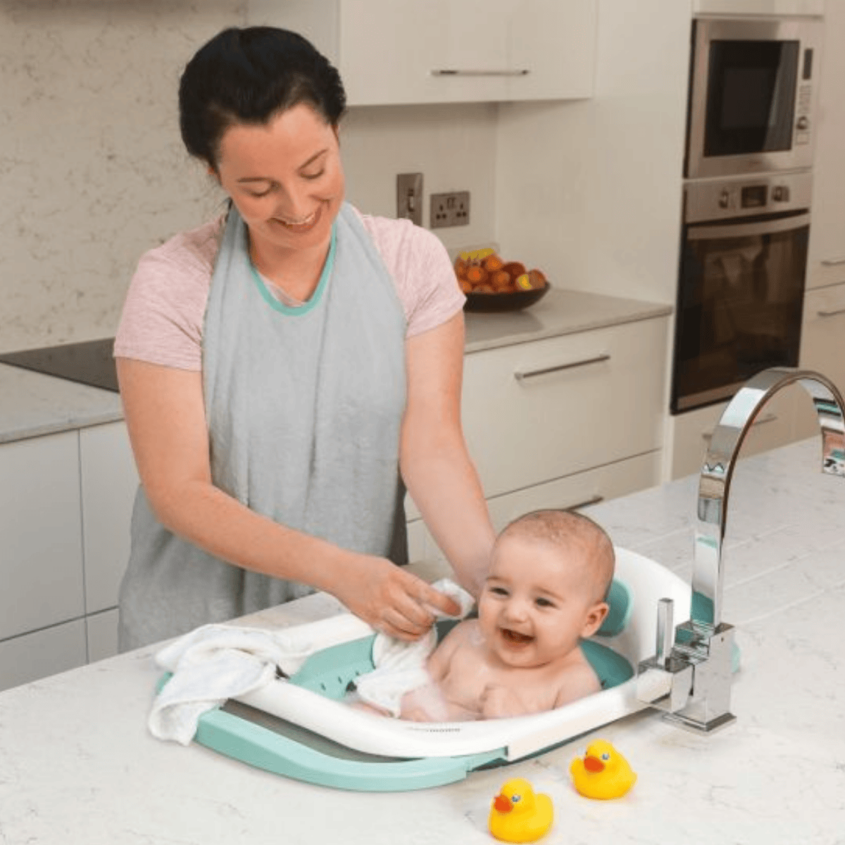 Woman bathing a baby in a kitchen using a baby bath seat with rubber ducks nearby.