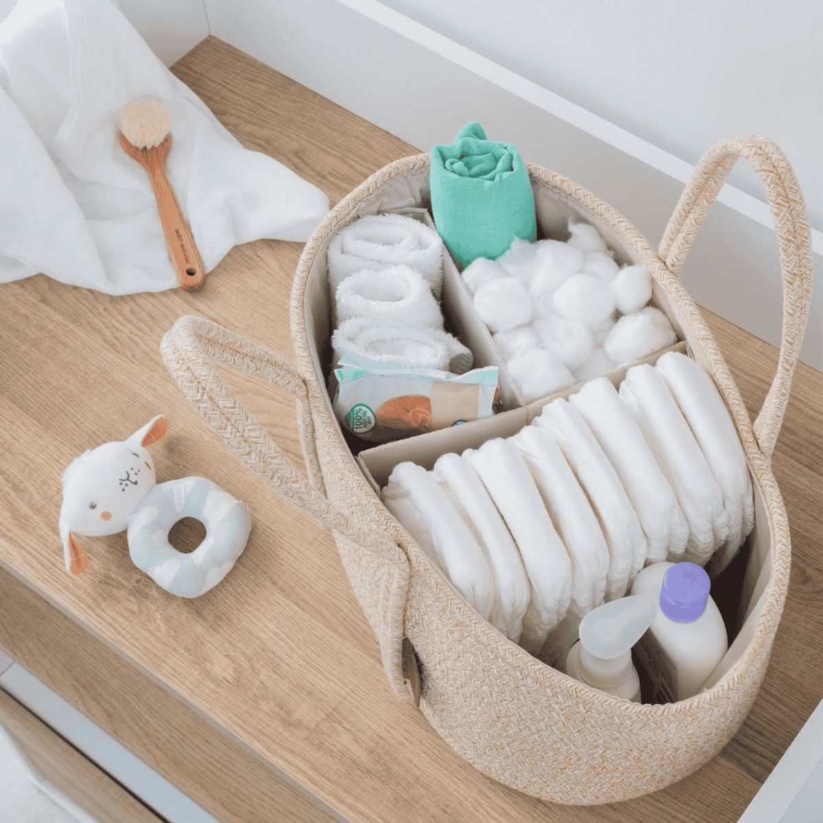 Beige storage basket with baby items including diapers, toys, and bottles on a wooden surface.