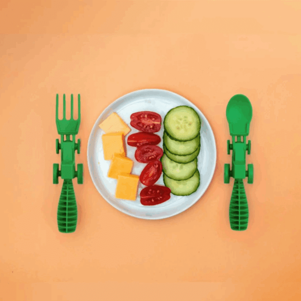 Children's green plastic fork and spoon on an orange background with a plate of sliced vegetables and cheese.