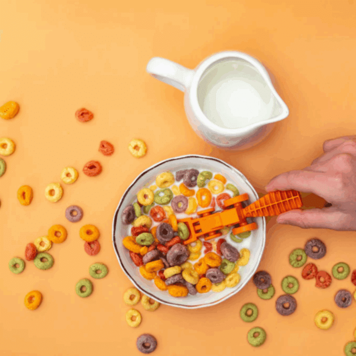 Colorful cereal in a bowl with a hand holding a small orange toy cutlery utensil on an orange background