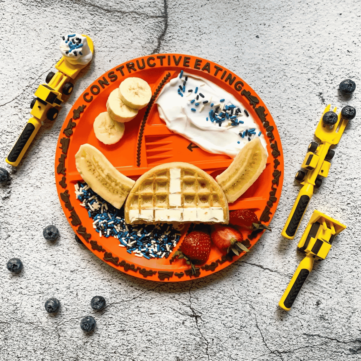 An overhead flat-lay of a children's waffle-themed plate on a textured gray surface, surrounded by toy construction tools. The orange plate has compartments with waffles, banana slices, strawberries, whipped cream, blue sprinkles, and the words 'CONSTRUCTIVE EATING'.