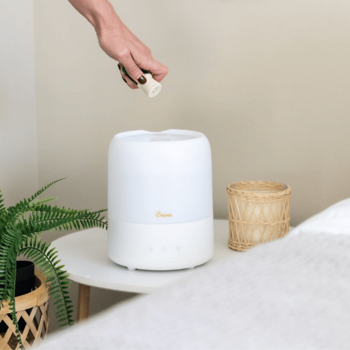 White humidifier on a table with a hand holding a bottle, a plant, and a basket in the background.