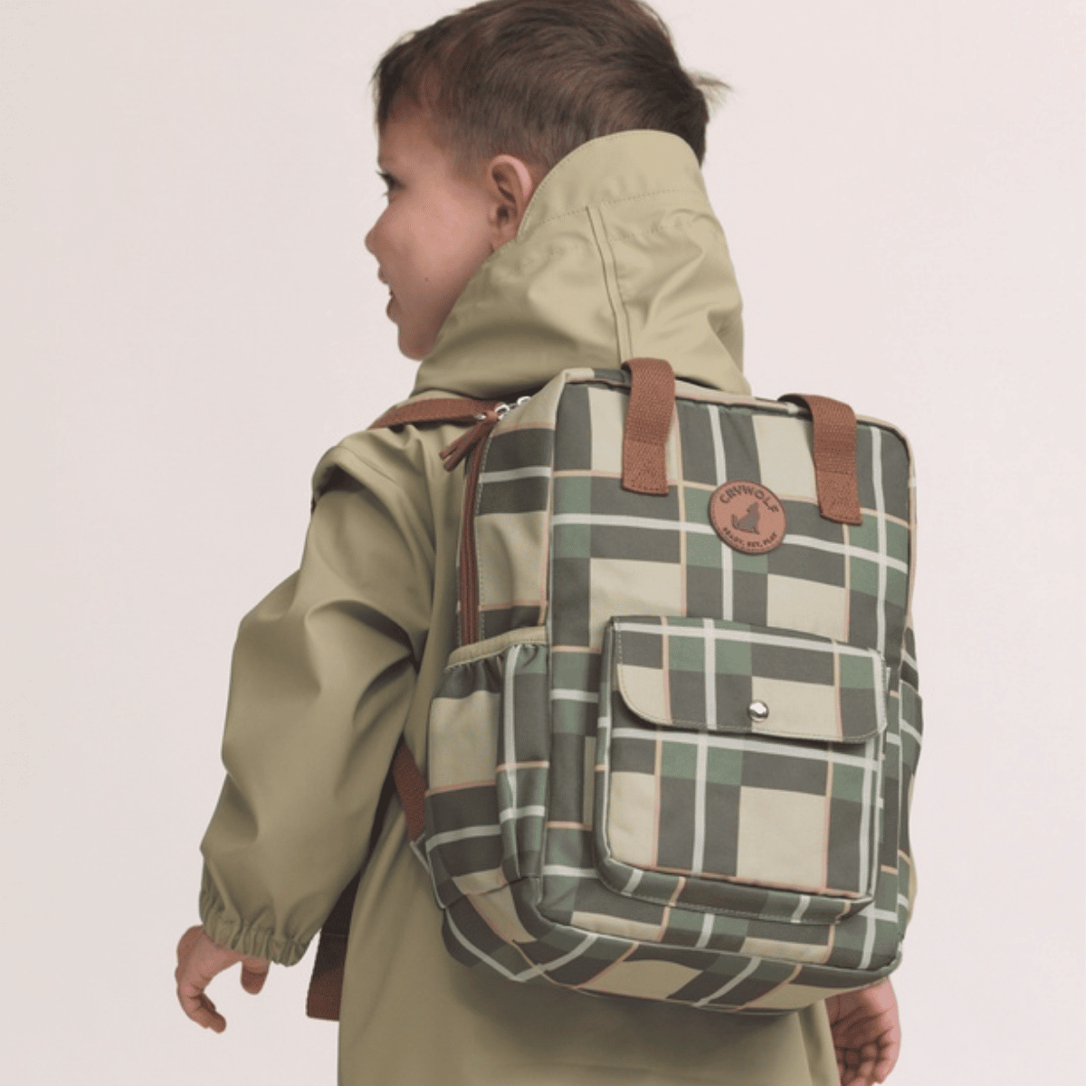 A child wearing an olive green hooded jacket is shown from the back, carrying a plaid backpack in shades of green, beige, and brown. The backpack features a front pocket with a snap button and a circular brown logo patch.