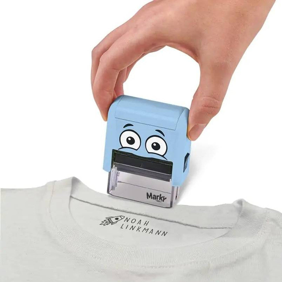 Hand using a blue cartoonish stamp on a white shirt with 'Noah Linkmann' logo.