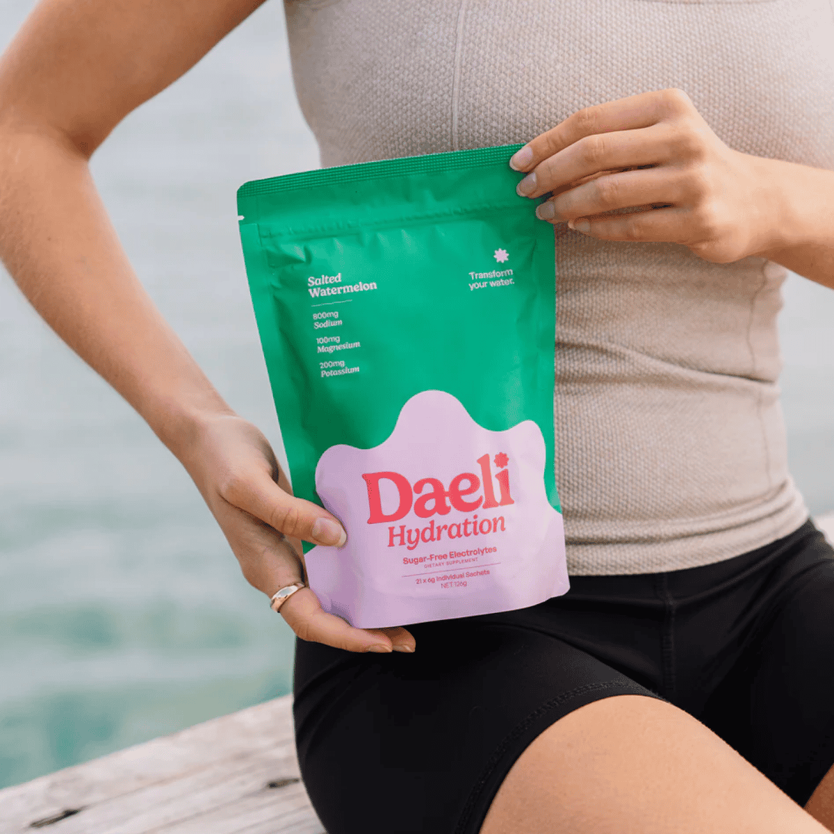 Person holding a green and pink Daeli Hydration package by a body of water.