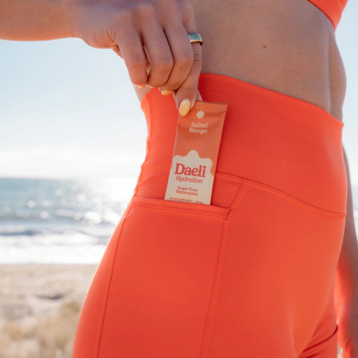 Person wearing bright orange shorts with a Daeli product in the pocket, beach background