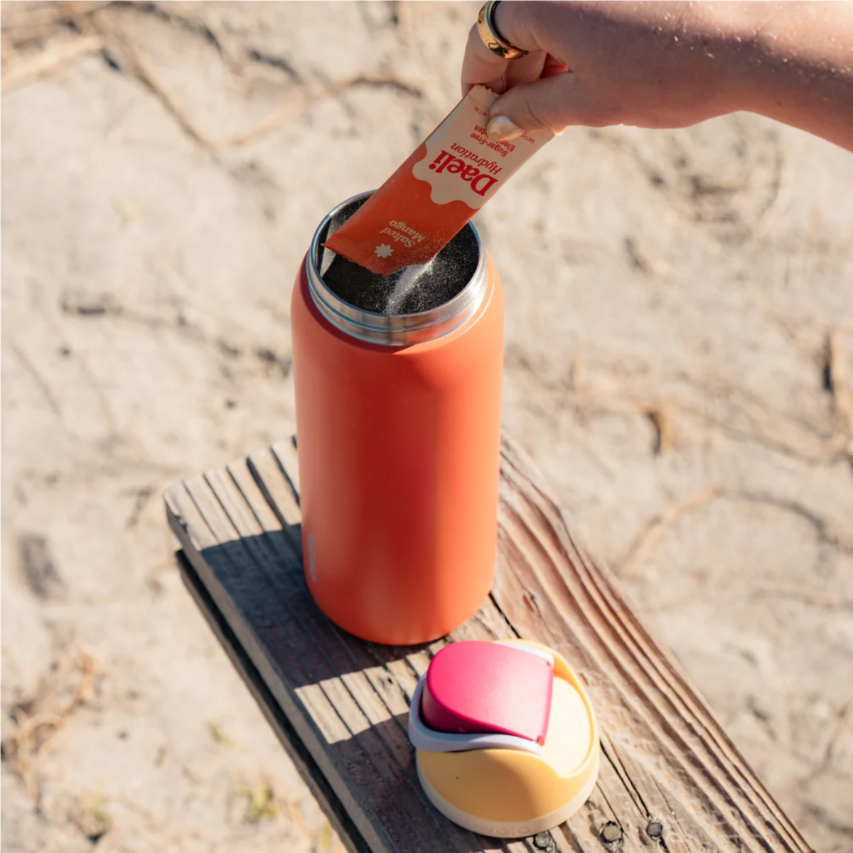 Person opening a red thermos with a straw on a sandy background