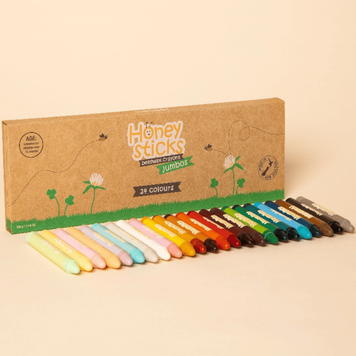 A set of 29 Honey Sticks Jumbos crayons in a variety of colors, arranged in a row in front of their brown kraft paper packaging box on a solid beige background.