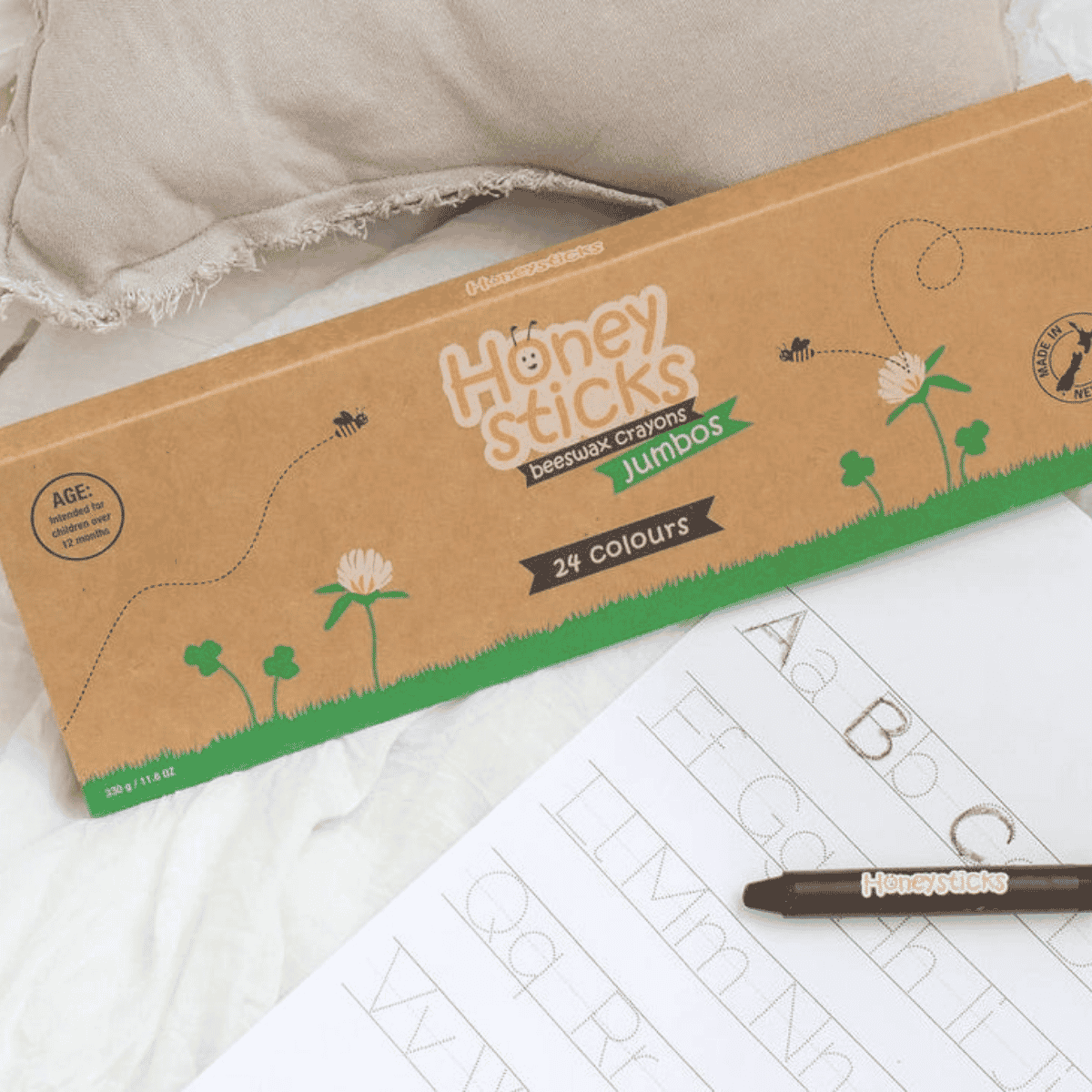 A box of Honey Sticks Jumbo crayons in 24 colours, styled on a white fabric surface next to a tracing sheet and a brown crayon.