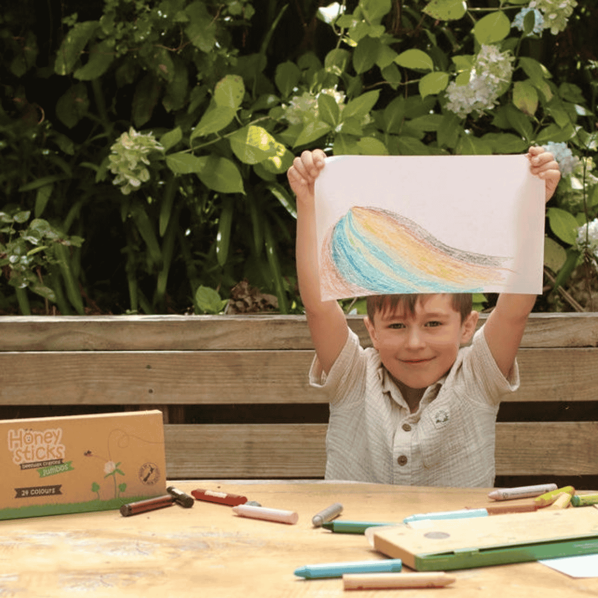A young boy sits at a wooden table outdoors, holding up a colored pencil drawing of a rainbow wave. A Honey Sticks crayon box is visible on the table next to other crayons and drawing supplies, with green garden foliage in the background.