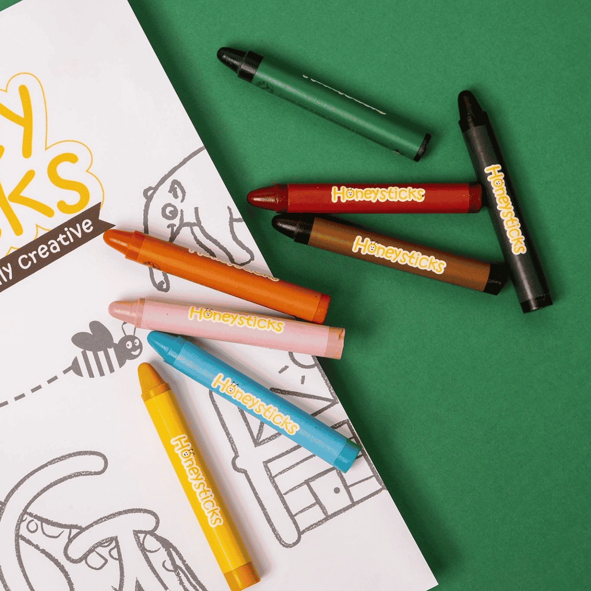 A set of Honeysticks crayons in multiple colors, including green, red, brown, black, orange, pink, blue, and yellow, arranged on a green background next to a white coloring page.
