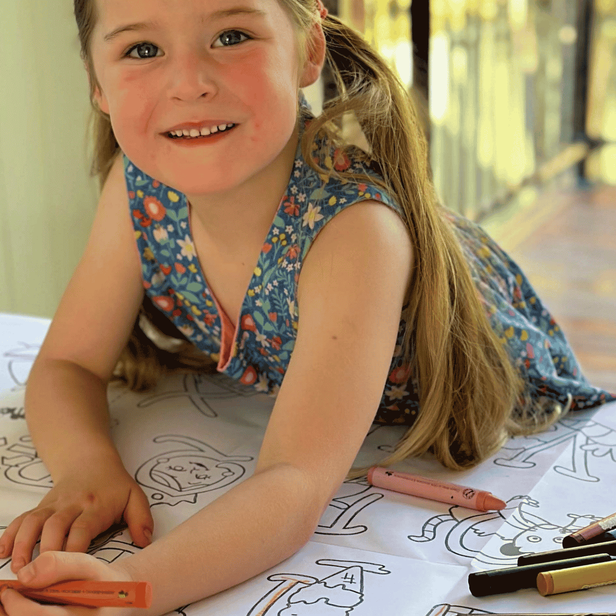 A young girl with blonde hair in a ponytail smiles while drawing on a large sheet of paper with crayons. She is wearing a sleeveless blue floral dress, and several crayons are scattered on the table.