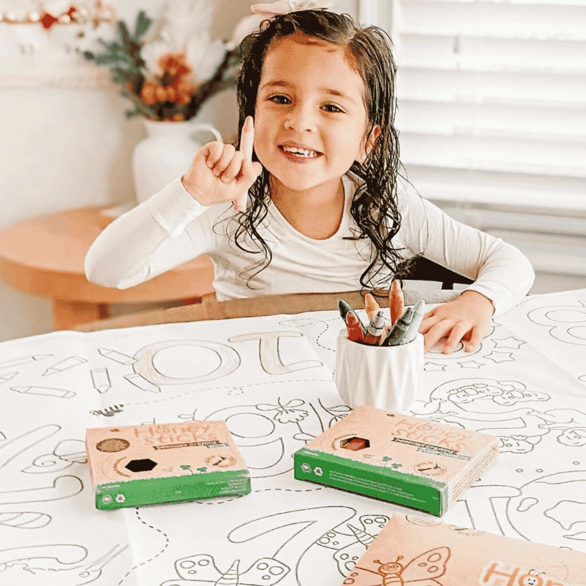 A young girl with curly hair sits at a table covered in a coloring book, smiling and pointing upward. Two 'Homer Simpson' themed coloring book boxes are visible on the table, along with a white cup holding colored pencils.