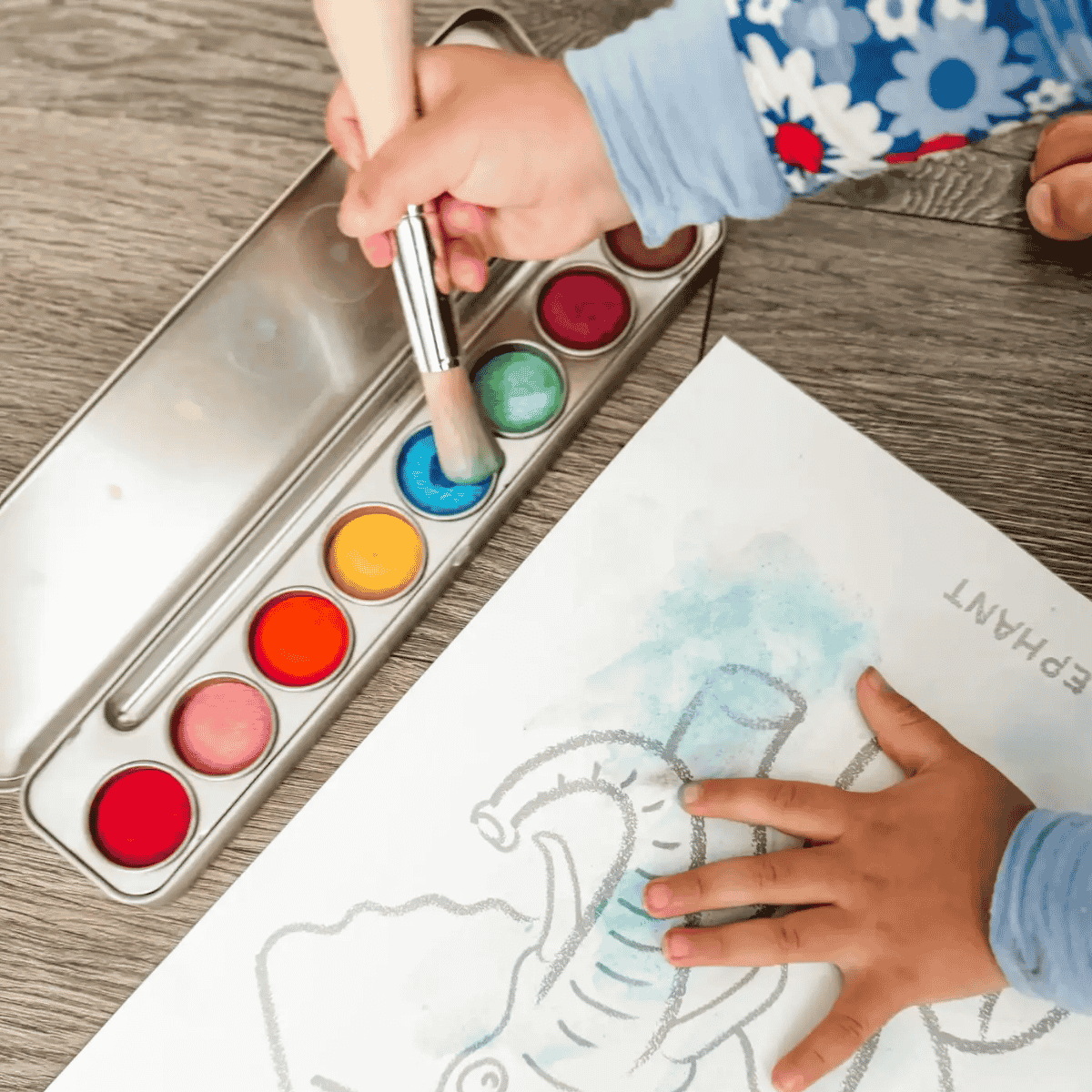 A close-up of a child's hands using a paintbrush to dip into a metal watercolor palette, with one hand painting on a white elephant drawing sheet. The palette contains multiple vibrant watercolor pans including blue, green, yellow, orange, red, and pink, set on a gray wood-grain tabletop.