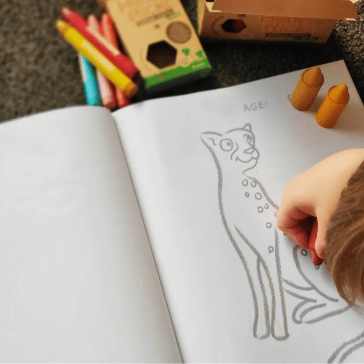 A child's hand coloring in a children's activity book with a cat outline, surrounded by crayons and wooden craft boxes on a dark textured surface.