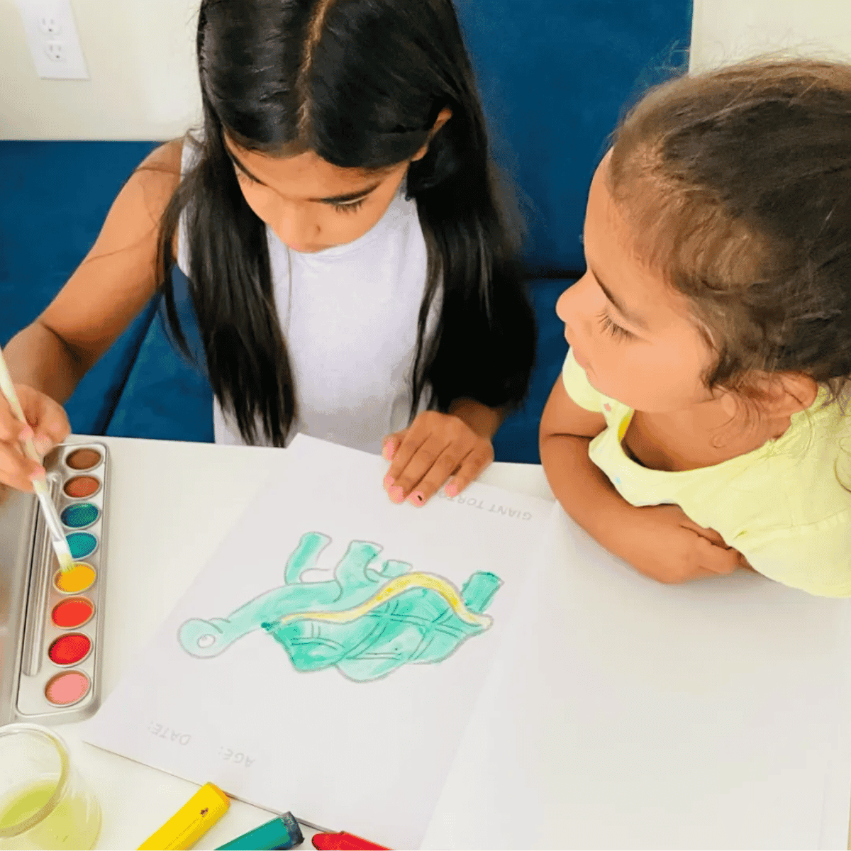 Two young girls sitting at a white table, one painting a green turtle with a yellow stripe on paper while the other looks on. A watercolor paint palette is visible on the left.