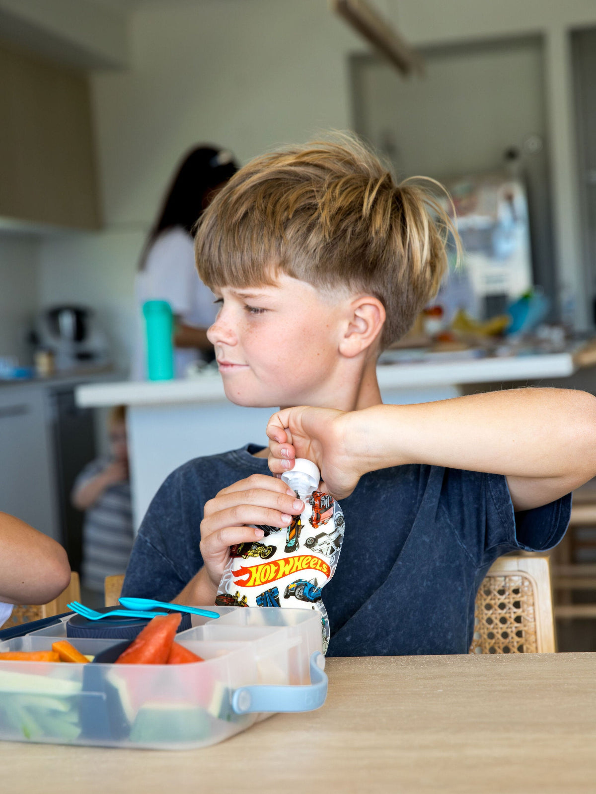 Child opening a Hot Wheels packaging at a table in a kitchen.