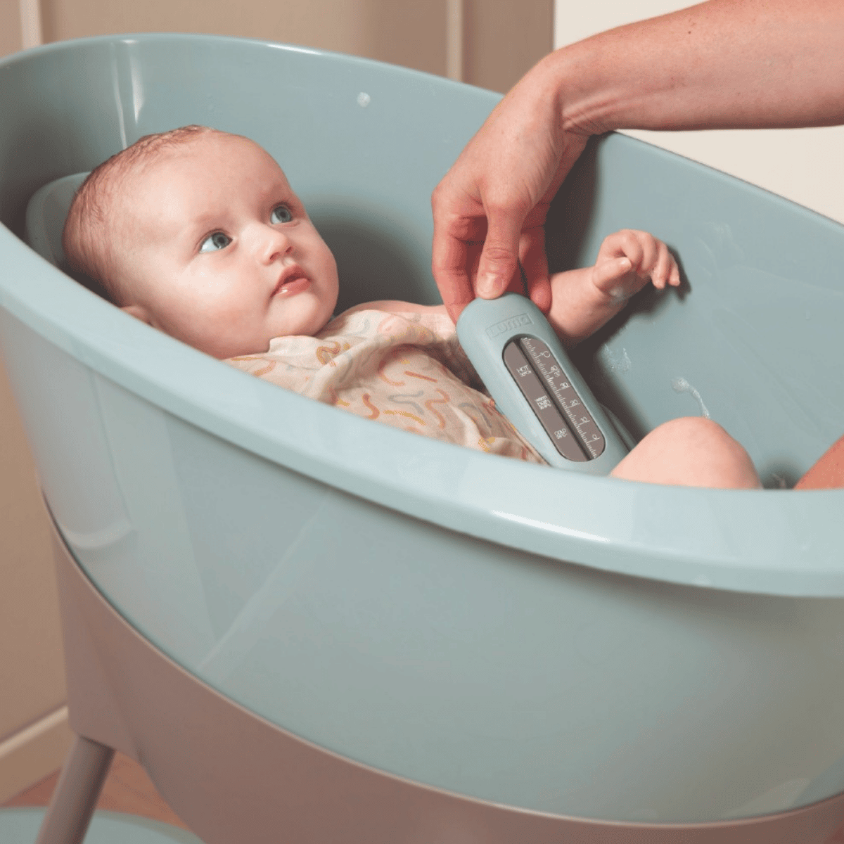 A baby sits in a light blue infant bath while an adult hand uses a matching light blue digital thermometer inside the tub.