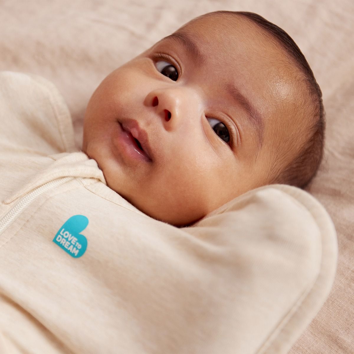 Baby wrapped in a beige swaddle with a blue 'Love & Dream' tag, lying on a soft surface.